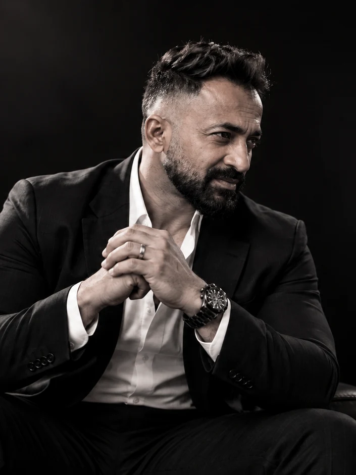 Satyajit Dasgupta seated in a dark suit, hands clasped, against a black ground.